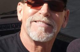 Photo of Dan Badgley in a black shirt, black hat with logo, and dark sunglasses. He is a white man with light gray facial hair, smiling at the camera in the sunshine.