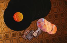 Placeholder image of vinyl records, cassette tapes, and CDs spread out on a yellow/brown background.