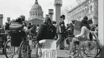 Black and white photograph of protestors in wheelchairs in San Francisco Civic Center with signs such as "We Shall Overcome" and "Equal Opportunity"