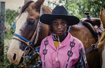 Photograph of African American cowboy wearing a pink shirt and black cowboy hat and standing in front of a horse