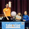 Man wearing orange shirt holds up bottle full of gas while kids look on in amazement. Table has a sign on it that says Talewise: Learning Powered by Science